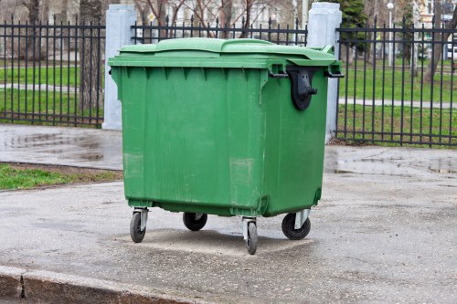 A large, green plastic wheelie bin with a closed lid, situated on a paved surface outdoors, likely used for rubbish collection. The bin has a textured surface with visible ridges along the top edge and is mounted on four small wheels, facilitating mobility. The background includes a black metal fence with vertical bars and concrete posts, a grassy area with trees, and a pathway or road. The overall scene suggests a typical rubbish disposal point in a residential or communal outdoor space, in an area accessible for waste collection services such as those provided by Commercial Waste Maidenhead, near Maidenhead town center. The lighting is natural, indicating daytime, and the environment appears clean and orderly, consistent with waste management environments in the UK.