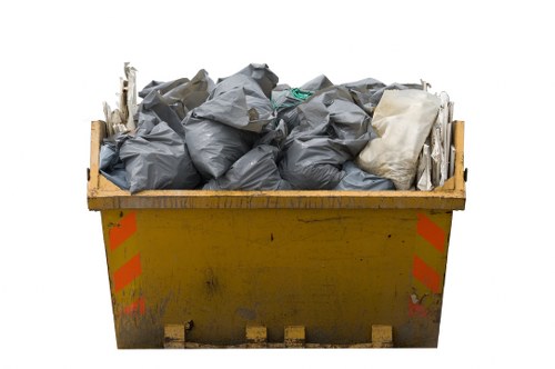 A large yellow industrial skip positioned on a paved surface, filled to the brim with black plastic rubbish bags and some off-white items, indicating waste collection for a commercial property in Maidenhead. The skip's sides are marked with faint orange reflective stripes, and the surface shows signs of dirt and usage. The mixture of tightly packed black rubbish bags suggests the waste is securely contained, while the off-white items, possibly cardboard or fabric, add to the varied contents. The scene is set outdoors, with the skip placed in a typical urban or industrial environment suitable for commercial waste collection, representing a task handled by services such as those provided by Commercial Waste Maidenhead, which specialises in rubbish removal in the Maidenhead area.