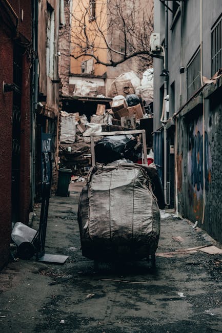 A narrow urban alleyway filled with accumulated rubbish and discarded items. In the foreground, a large, grey, weathered trash bag is set on a trolley with metal handles, positioned on a dark, gritty 