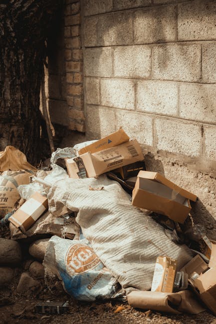 A narrow urban alleyway filled with accumulated rubbish and discarded items. In the foreground, a large, grey, weathered trash bag is set on a trolley with metal handles, positioned on a dark, gritty 