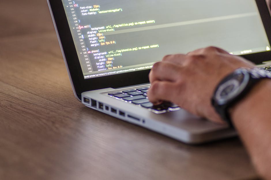 A close-up photograph of a person's hand typing on a silver laptop keyboard placed on a wooden surface. The person's wrist is adorned with a black watch featuring a round face and multiple dials. The 