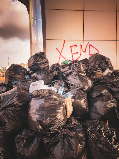 A narrow urban alleyway filled with accumulated rubbish and discarded waste materials. In the foreground, there is a large, grey, weathered garbage bag or sack positioned on the asphalt ground, which 